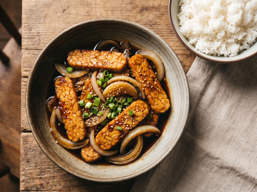 Tempeh acebolado dourado em prato de cerâmica, com cebolas caramelizadas, molho de shoyu, cebolinha e gergelim, servido com arroz branco