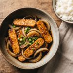 Tempeh acebolado dourado em prato de cerâmica, com cebolas caramelizadas, molho de shoyu, cebolinha e gergelim, servido com arroz branco