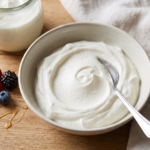 Iogurte grego vegano cremoso e denso em tigela de cerâmica clara, com marca da colher em relevo, servido com frutas vermelhas
