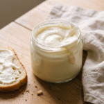Pote de vidro com requeijão vegano cremoso branco e espesso, ao lado de uma fatia de pão com requeijão passado