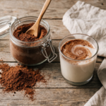 Pote de vidro com achocolatado em pó caseiro e colher enfiada, ao lado de um copo de leite vegetal com o pó se dissolvendo