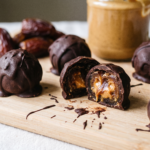 Bombons de tâmara cobertos de chocolate sobre tábua de madeira, um partido ao meio mostrando o recheio de pasta de amendoim