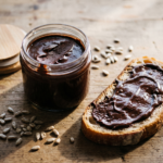 Pote de vidro com creme de chocolate escuro e brilhante, fatia de pão com o creme passado e sementes de girassol na mesa de madeira